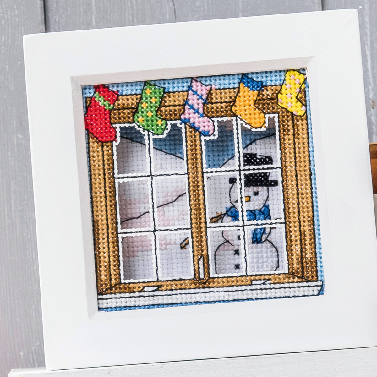 Cross-stitched scene of a window with snowmen and stockings, framed on a white background.