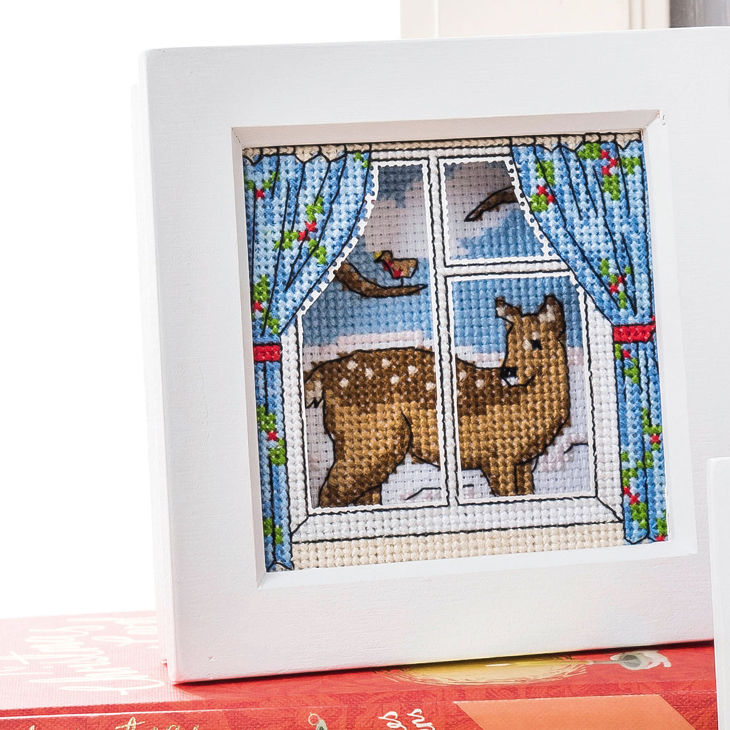 Cross-stitched deer looking out a window with blue curtains and a bird.