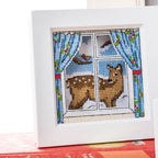 Cross-stitched deer looking out a window with blue curtains and a bird.