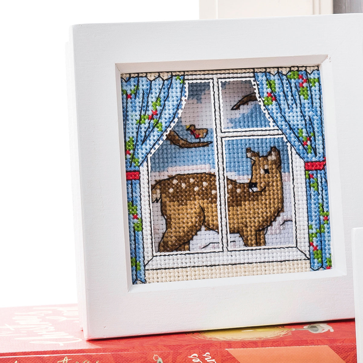 Cross-stitched deer looking out a window with blue curtains and a bird.