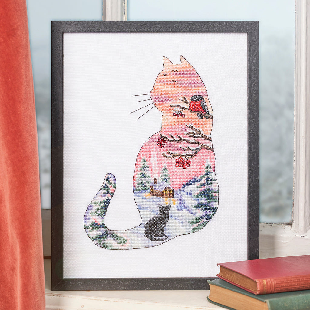 Framed cross-stitch art of a cat with a winter scene inside, on a light-colored wall.