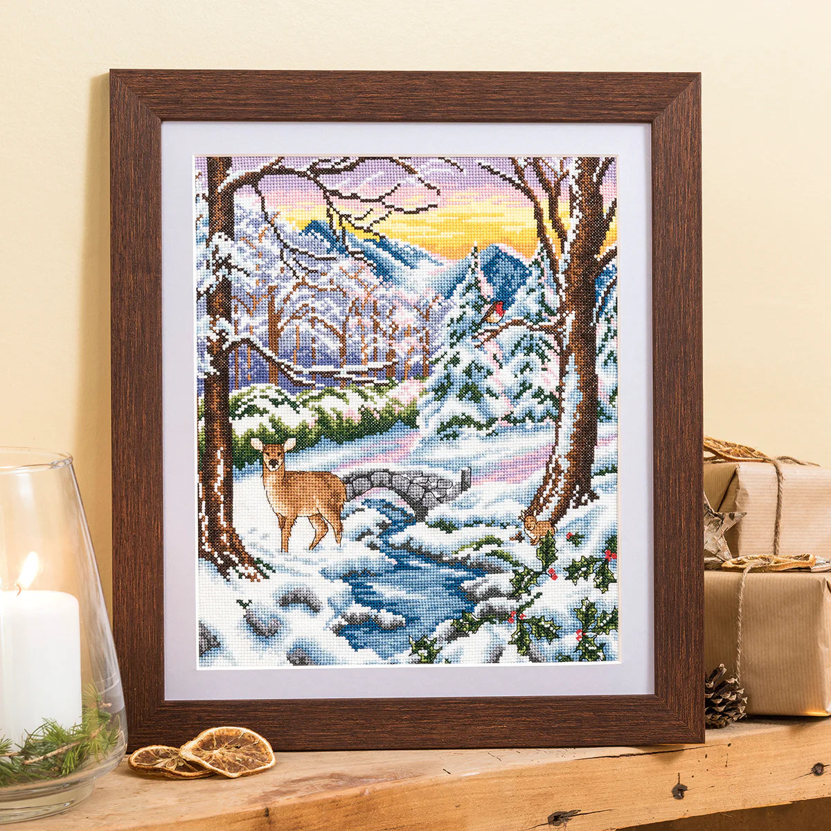 Framed cross-stitch art of a winter scene with a deer by a stream on a wooden surface.