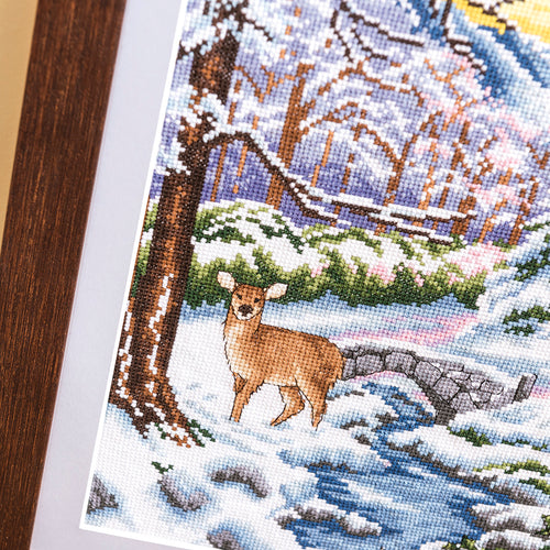Cross-stitched deer in a snowy landscape with trees and a bridge.