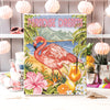 Cross-stitched picture of a flamingo with 'Paradise Dreams' text on a table with decorative items.