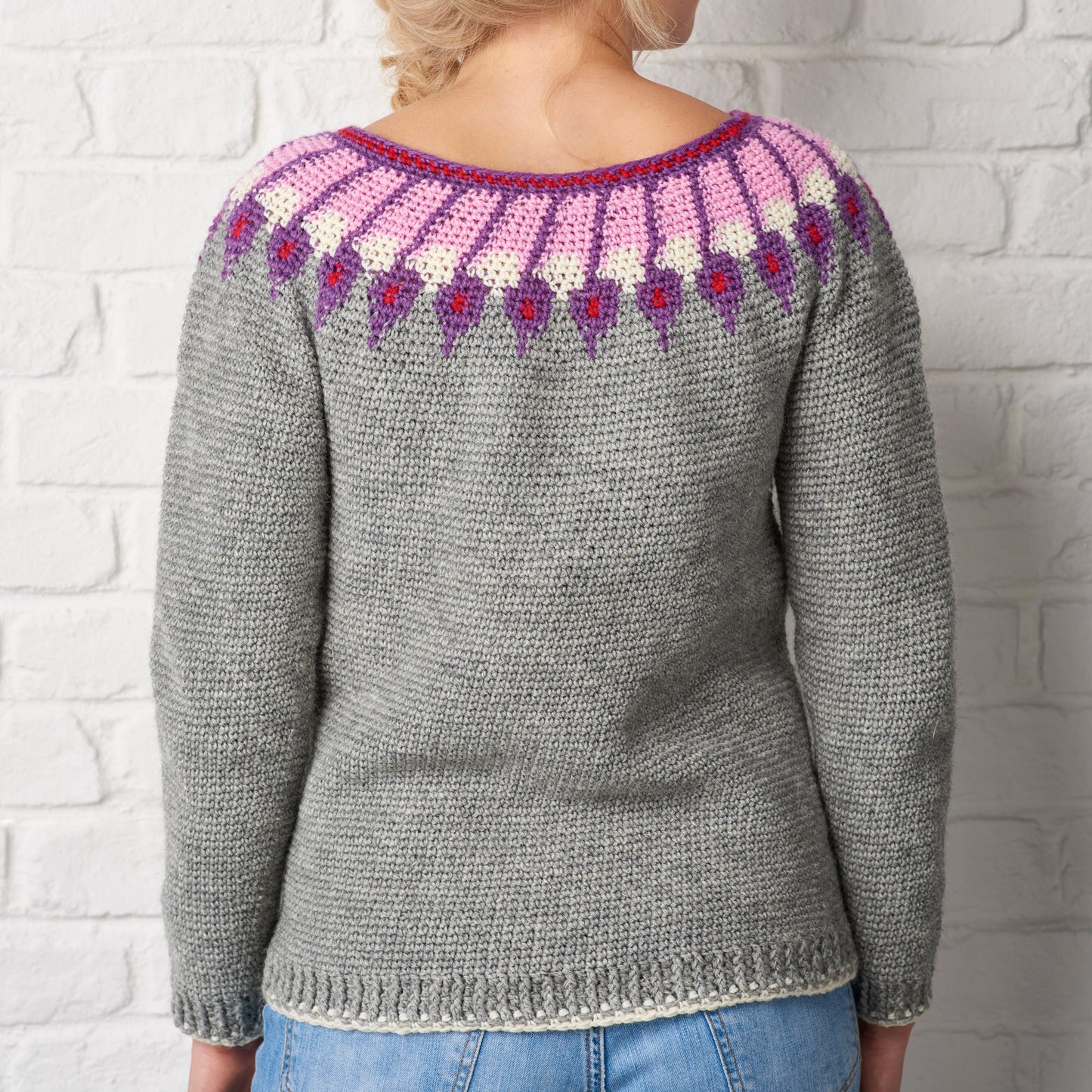 A person in a gray knitted sweater with colorful pink, purple, and white patterns on the yoke. They stand against a white brick wall. Cozy and casual vibe.