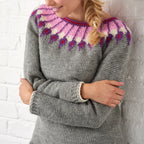 Person with braided hair wearing a cozy gray sweater featuring a vibrant pink, purple, and white patterned yoke, standing against a white brick wall.