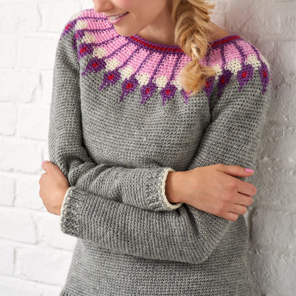 Person with braided hair wearing a cozy gray sweater featuring a vibrant pink, purple, and white patterned yoke, standing against a white brick wall.