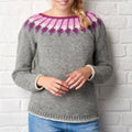 A woman wearing a gray knitted sweater with a colorful pink and purple patterned neckline stands against a white brick wall, hands gently clasped.