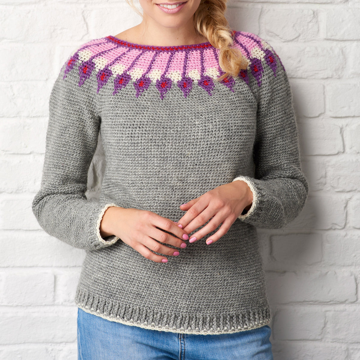 A woman wearing a gray knitted sweater with a colorful pink and purple patterned neckline stands against a white brick wall, hands gently clasped.