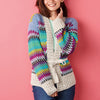 Smiling woman wearing a colorful crocheted cardigan with zigzag patterns in blue, purple, and yellow hues, against a pink background. Cozy and cheerful vibe.