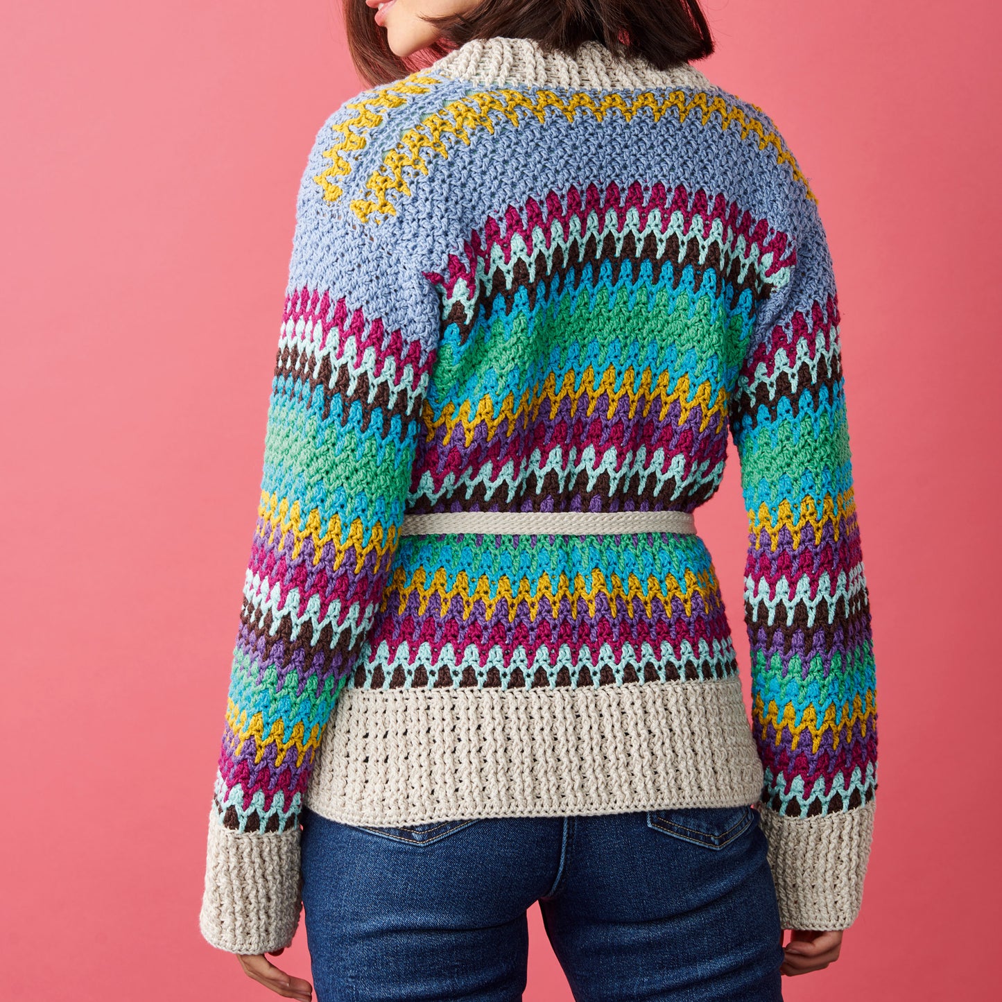 Woman in jeans wearing a colorful, knitted sweater with vibrant patterns stands against a pink background, conveying a cozy and cheerful vibe.
