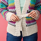 A person ties the belt of a colorful, textured cardigan with multicolored zigzag patterns over a mint green top. The background is pink.