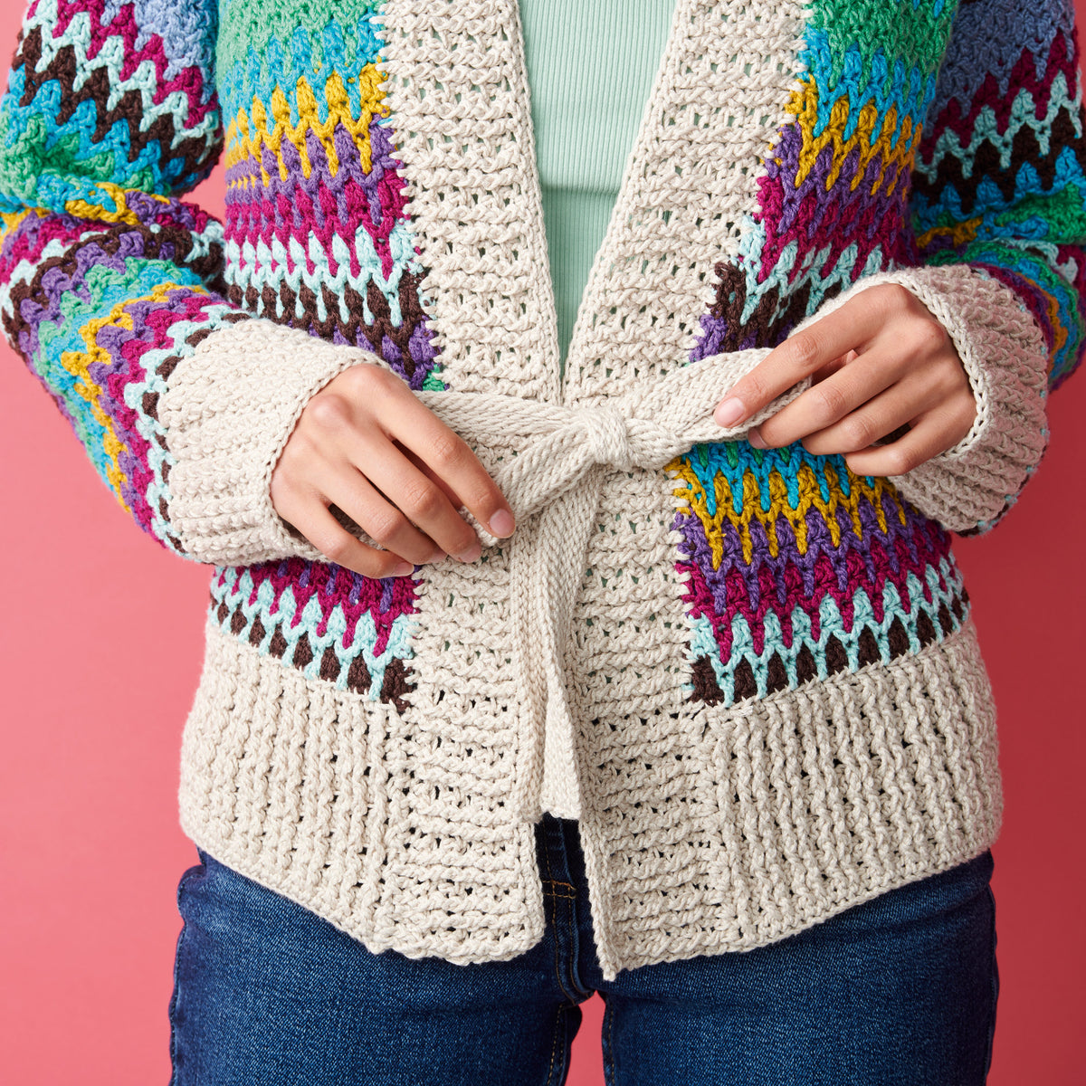 A person ties the belt of a colorful, textured cardigan with multicolored zigzag patterns over a mint green top. The background is pink.