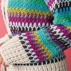 Close-up of a colorful, textured knit sweater with zigzag patterns in shades of pink, blue, yellow, and brown, set against a pink background.