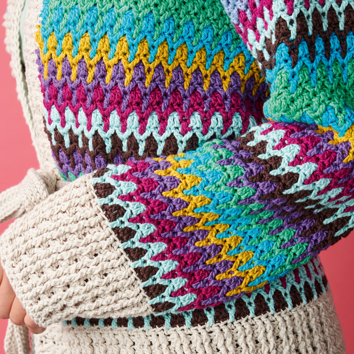 Close-up of a colorful, textured knit sweater with zigzag patterns in shades of pink, blue, yellow, and brown, set against a pink background.