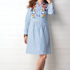 Woman wearing a blue striped dress with floral embroidery against a white wooden background