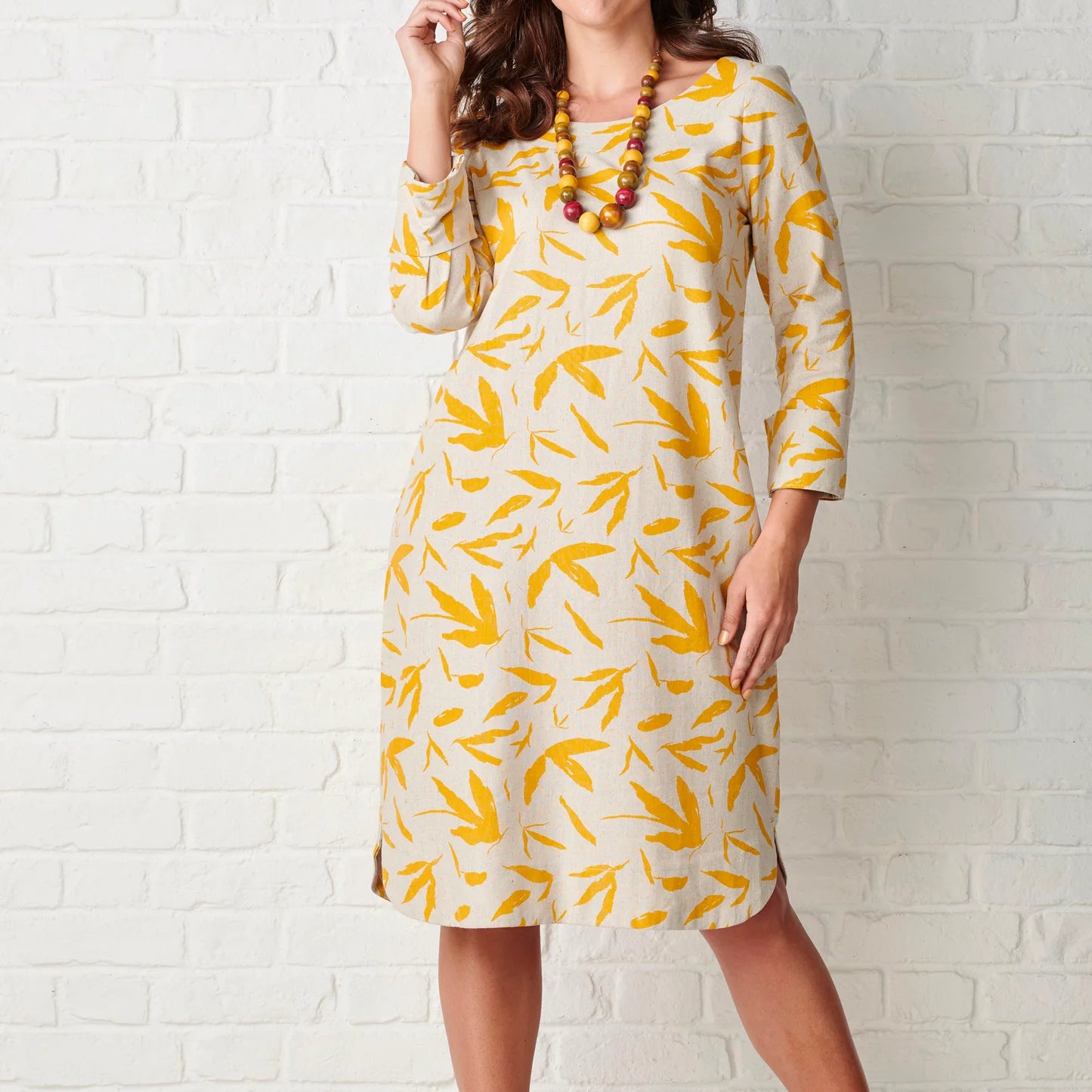Woman wearing a yellow and white patterned dress against a white brick wall.