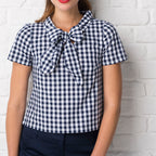 Woman wearing a blue and white checkered blouse with a bow tie against a white brick wall.