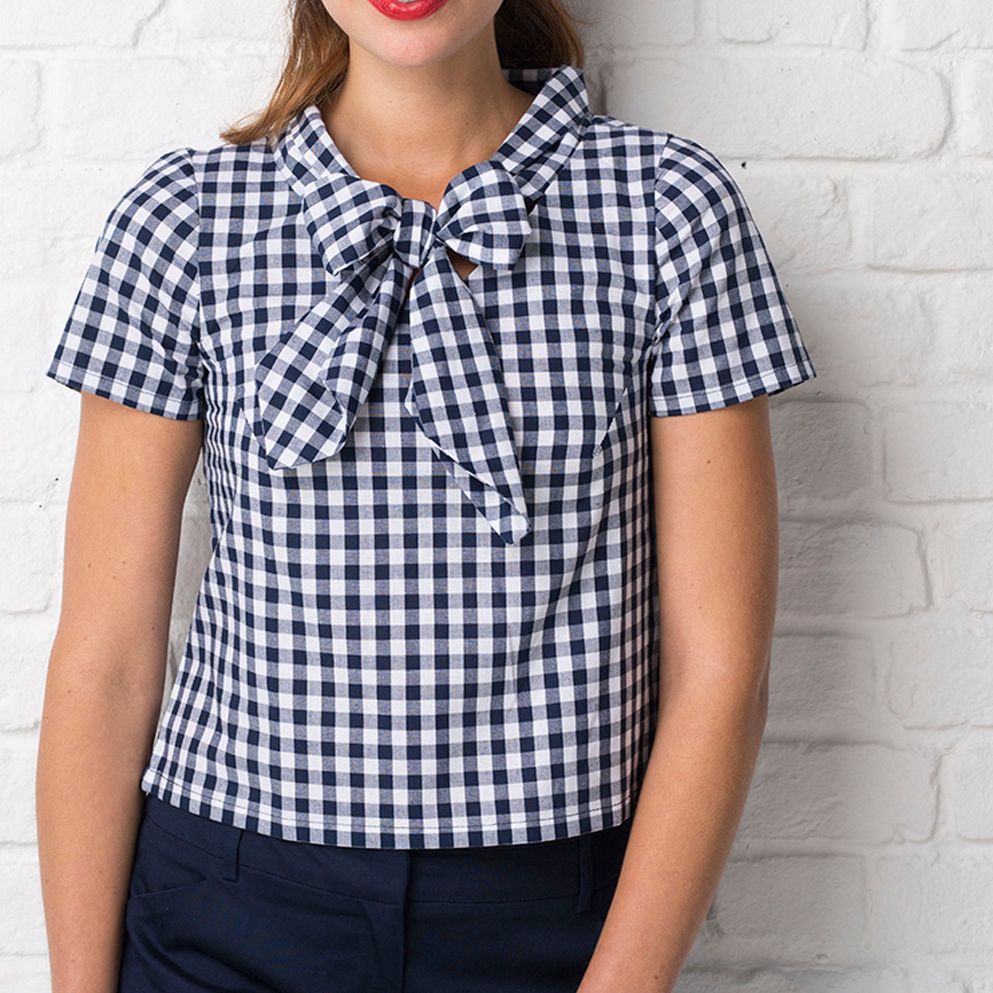 Woman wearing a blue and white checkered blouse with a bow tie against a white brick wall.