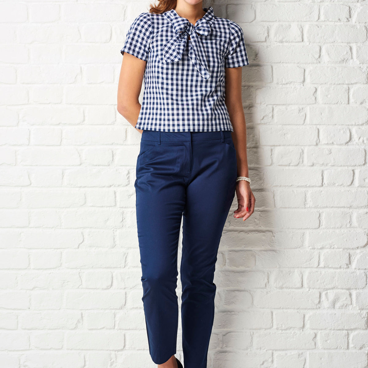 Person wearing a blue and white checkered shirt and navy pants against a white brick wall.