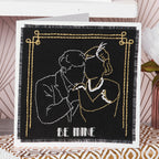 Greeting card with embroidered design of two people embracing and 'BE MINE' text on a black background.