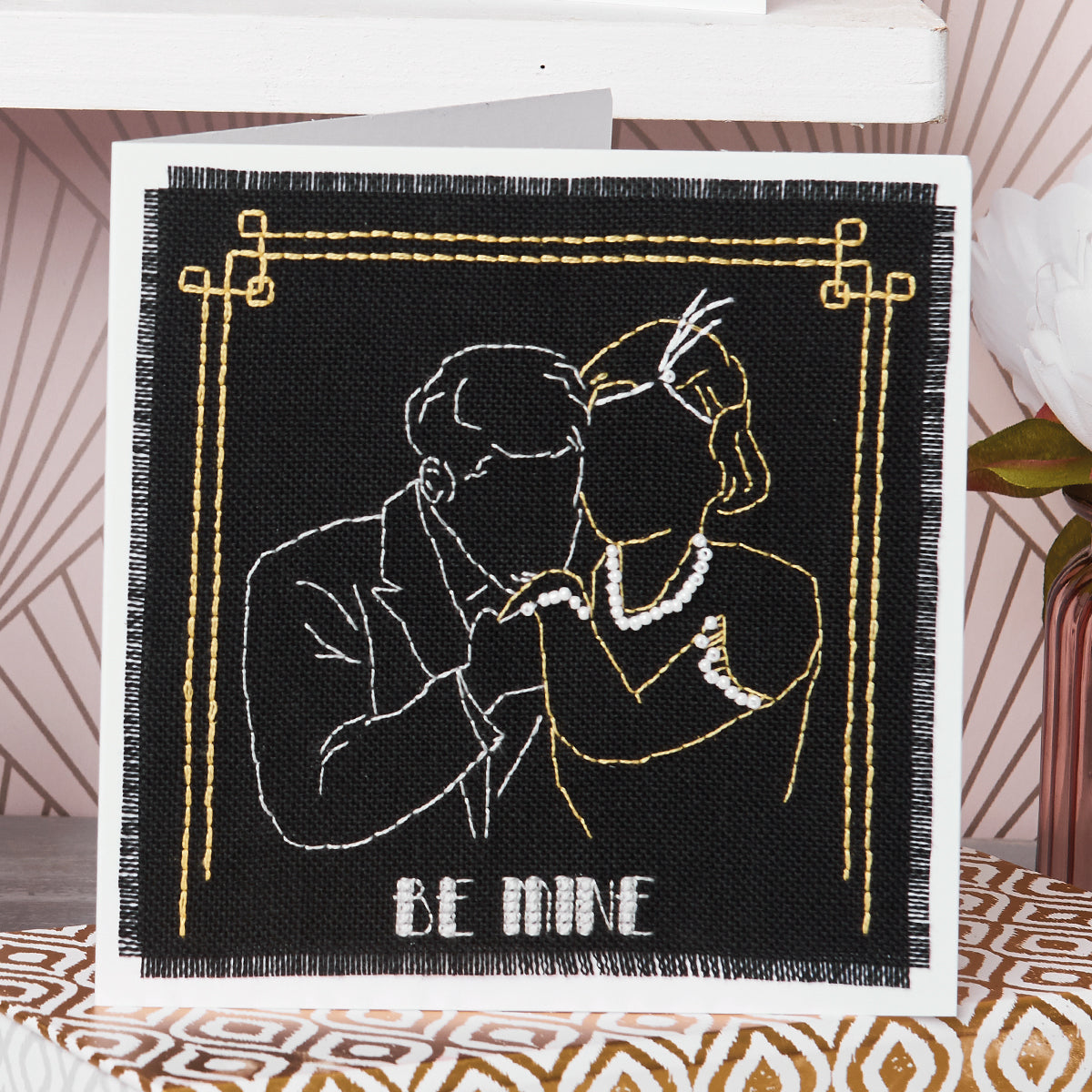 Greeting card with embroidered design of two people embracing and 'BE MINE' text on a black background.