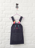 Children's denim pinafore dress with cat face design on a white brick wall background.