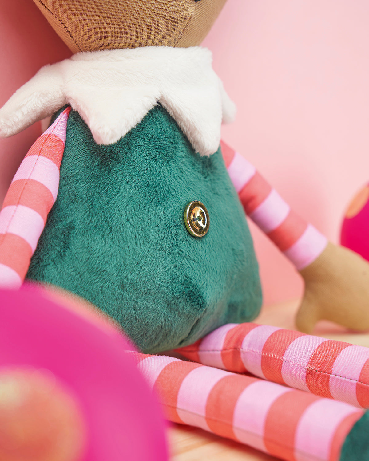 A plush toy with a green fuzzy body and white collar sits against a pink background. Its arms and legs are striped in pink and red, with a gold button detail.