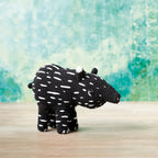 A crocheted black toy animal resembling a tapir stands on a wooden surface against a textured teal background. The toy has white spots and a playful appearance.