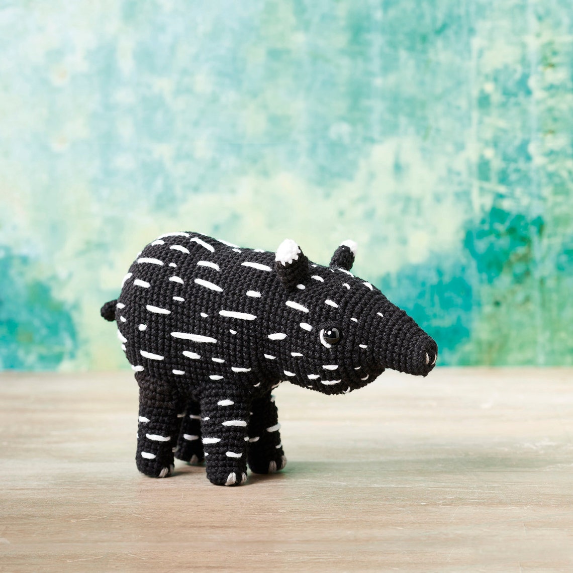 A crocheted black toy animal resembling a tapir stands on a wooden surface against a textured teal background. The toy has white spots and a playful appearance.