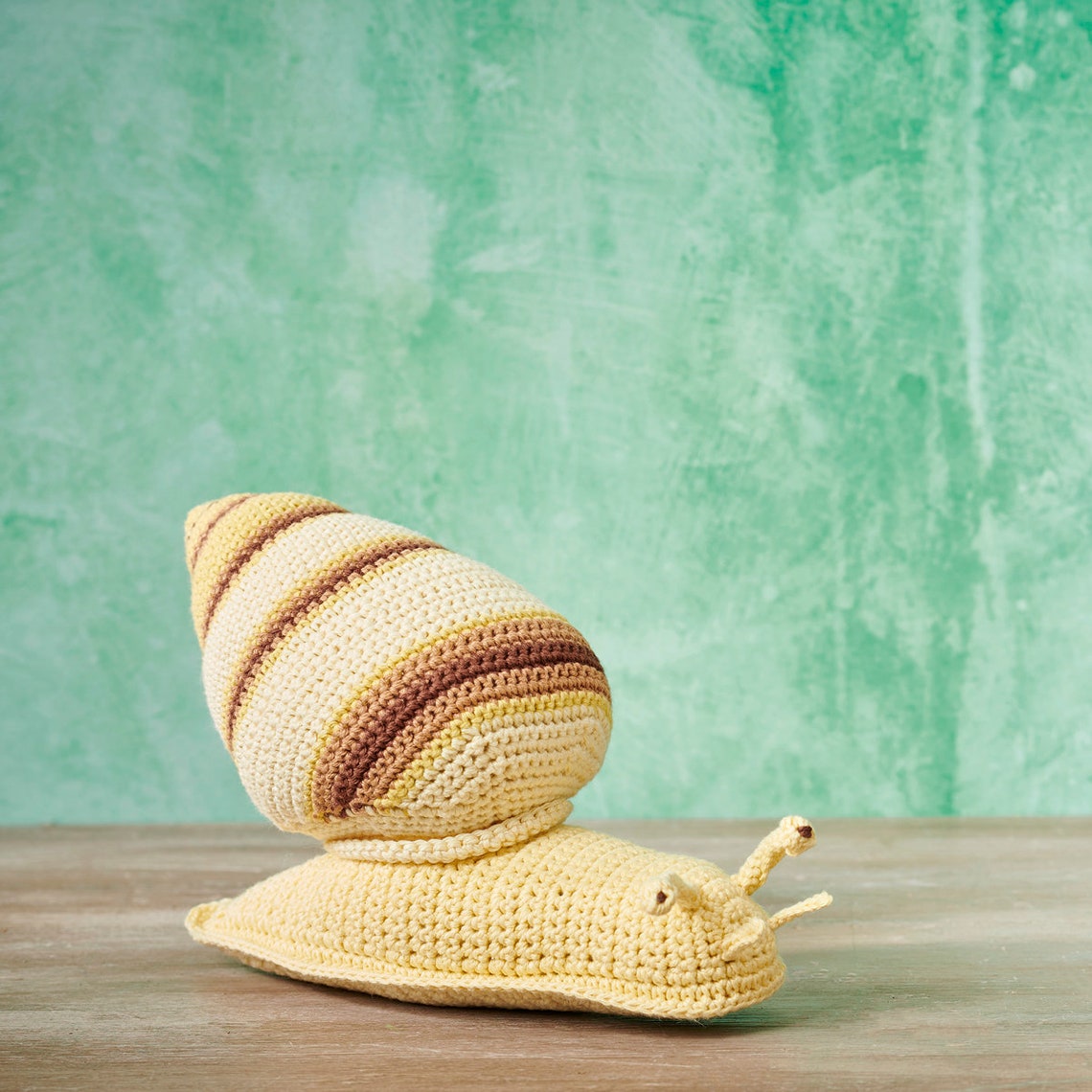 Crocheted snail toy with a yellow and brown striped shell on a wooden surface. The background is a textured, light green wall.