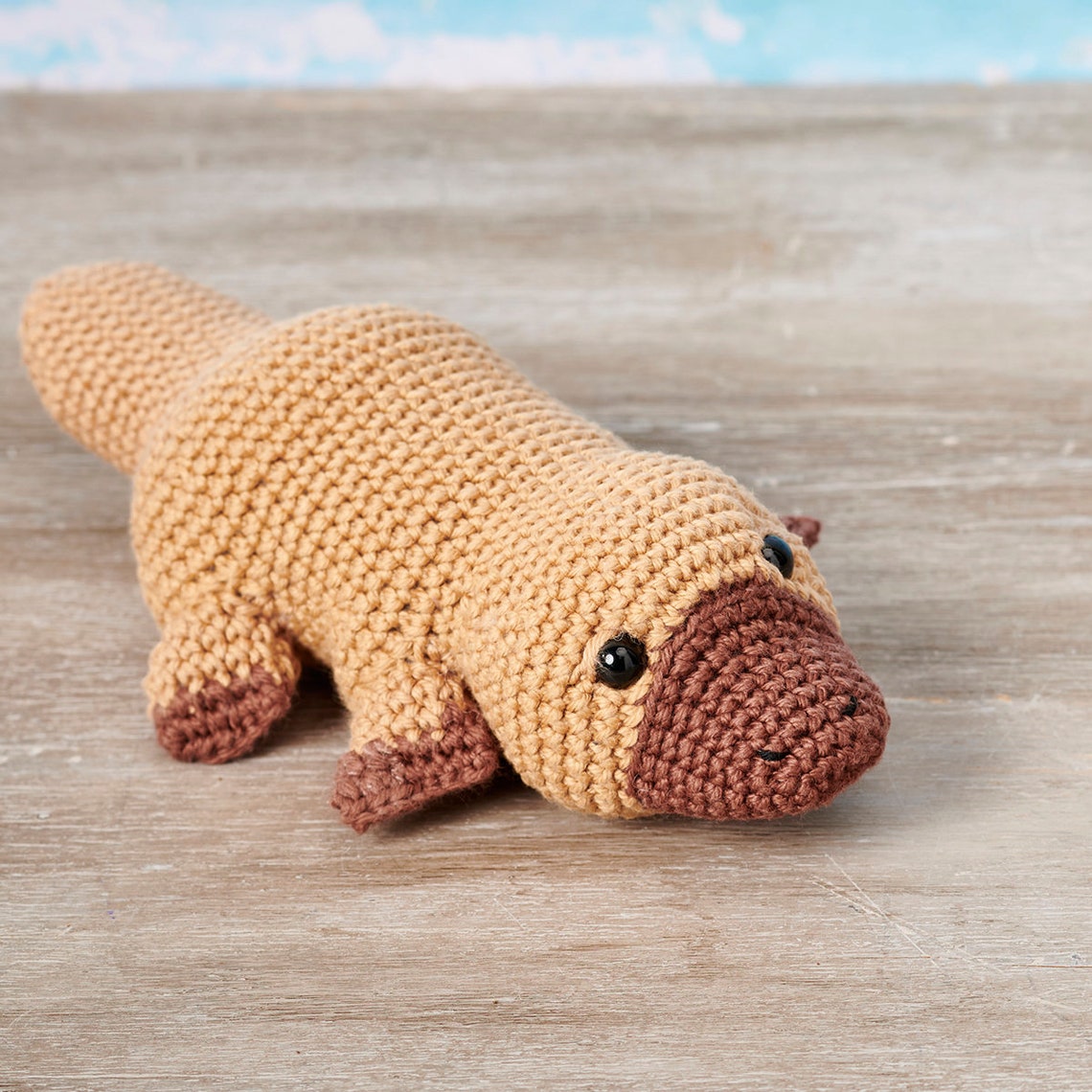 A crocheted platypus toy with a tan body and brown beak and feet lies on a wooden surface. It has black button eyes, conveying a cute and playful vibe.