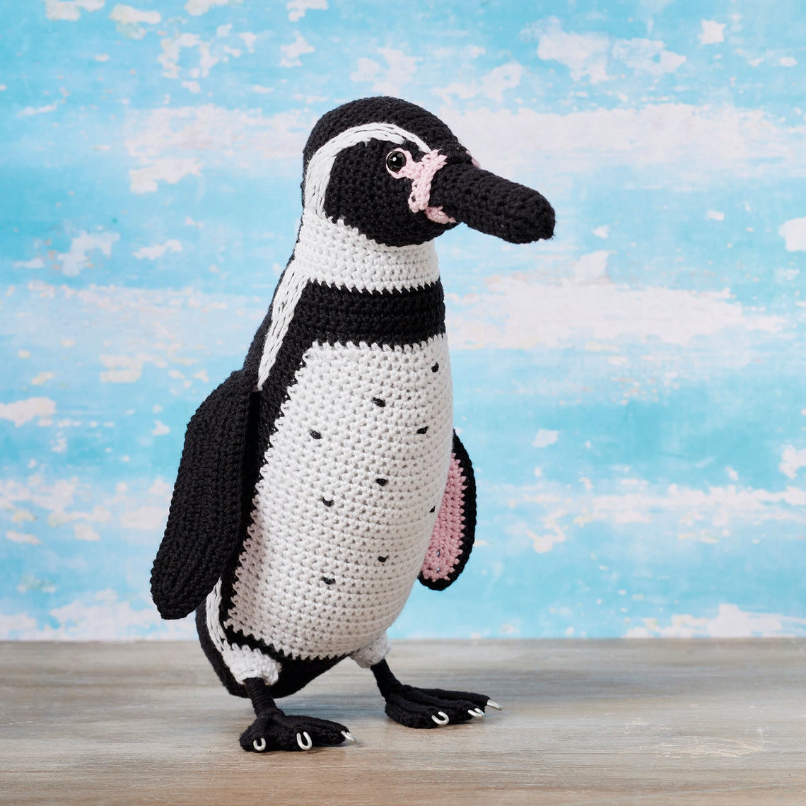 Crocheted penguin toy stands on a wooden surface against a blue sky background, featuring black, white, and pink yarn details, conveying a playful and cozy vibe.
