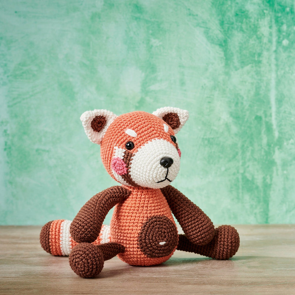 Crocheted red panda toy with orange, brown, and white details sits on a wooden surface. The background is a soft green, creating a playful tone.