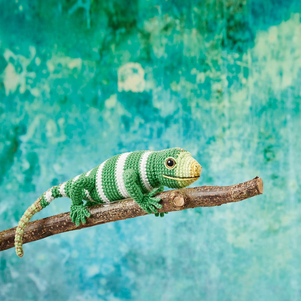 Crocheted green and white lizard perched on a branch, against a vibrant turquoise background. The scene feels playful and creative.