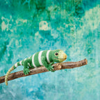 Crocheted green and white lizard perched on a branch, against a vibrant turquoise background. The scene feels playful and creative.