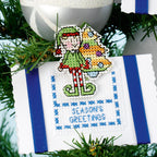 Cross-stitched ornament with an elf and Christmas tree design on a white background with blue stripes, surrounded by greenery.