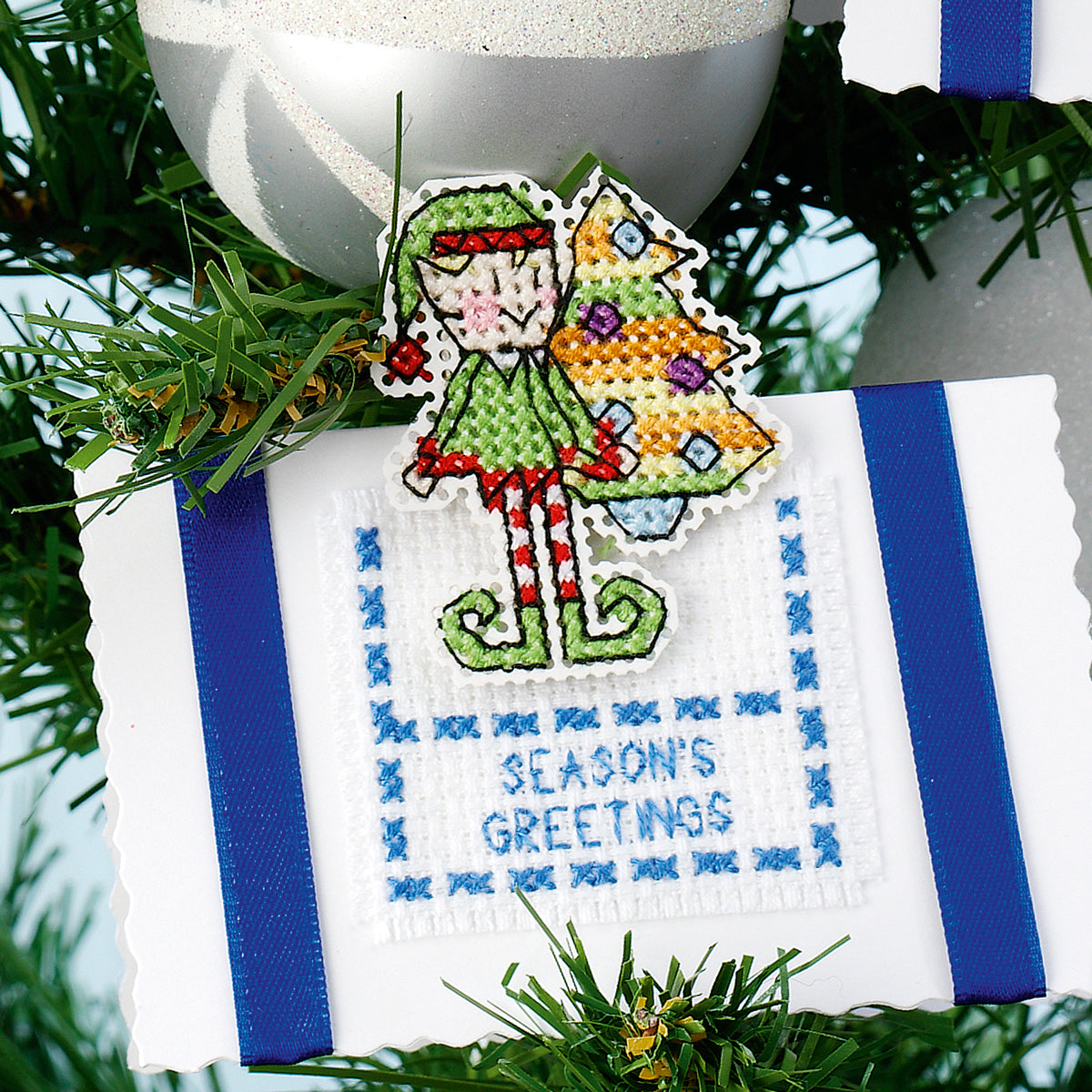 Cross-stitched ornament with an elf and Christmas tree design on a white background with blue stripes, surrounded by greenery.