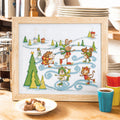 Framed cross-stitched picture of animals skiing on a snowy landscape, placed on a table with a cup and cookies.