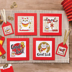 Set of cross-stitch patterns featuring lucky cats and 'Good Luck' text on a wooden surface with decorative elements.