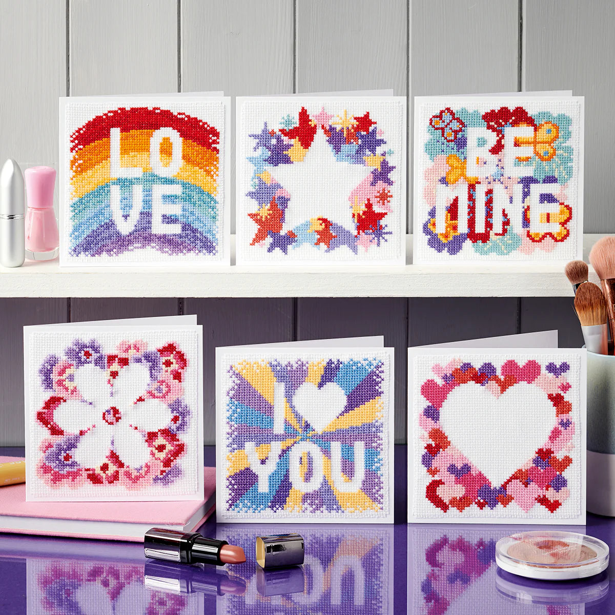 Set of cross-stitched cards with colorful designs on a gray wooden surface.