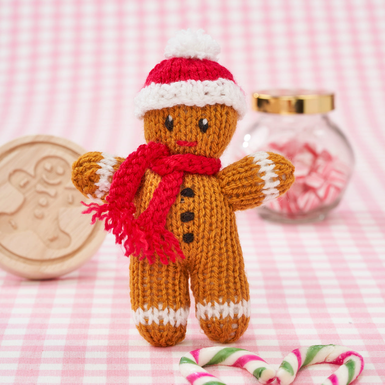 Knitted gingerbread man toy with a red scarf on a pink checkered background