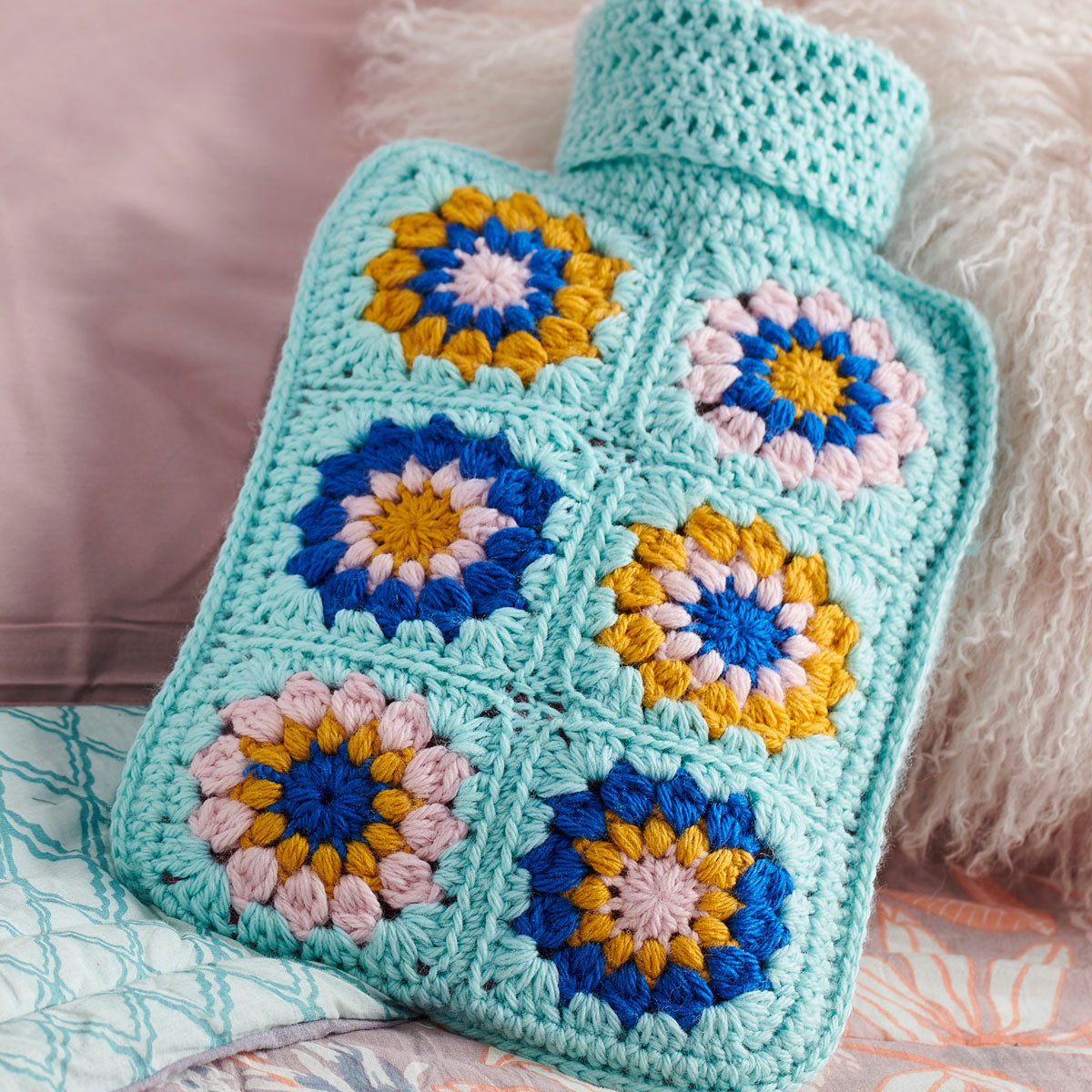 A crochet hot water bottle cover with a floral granny square pattern in blue, yellow, and pink colors.