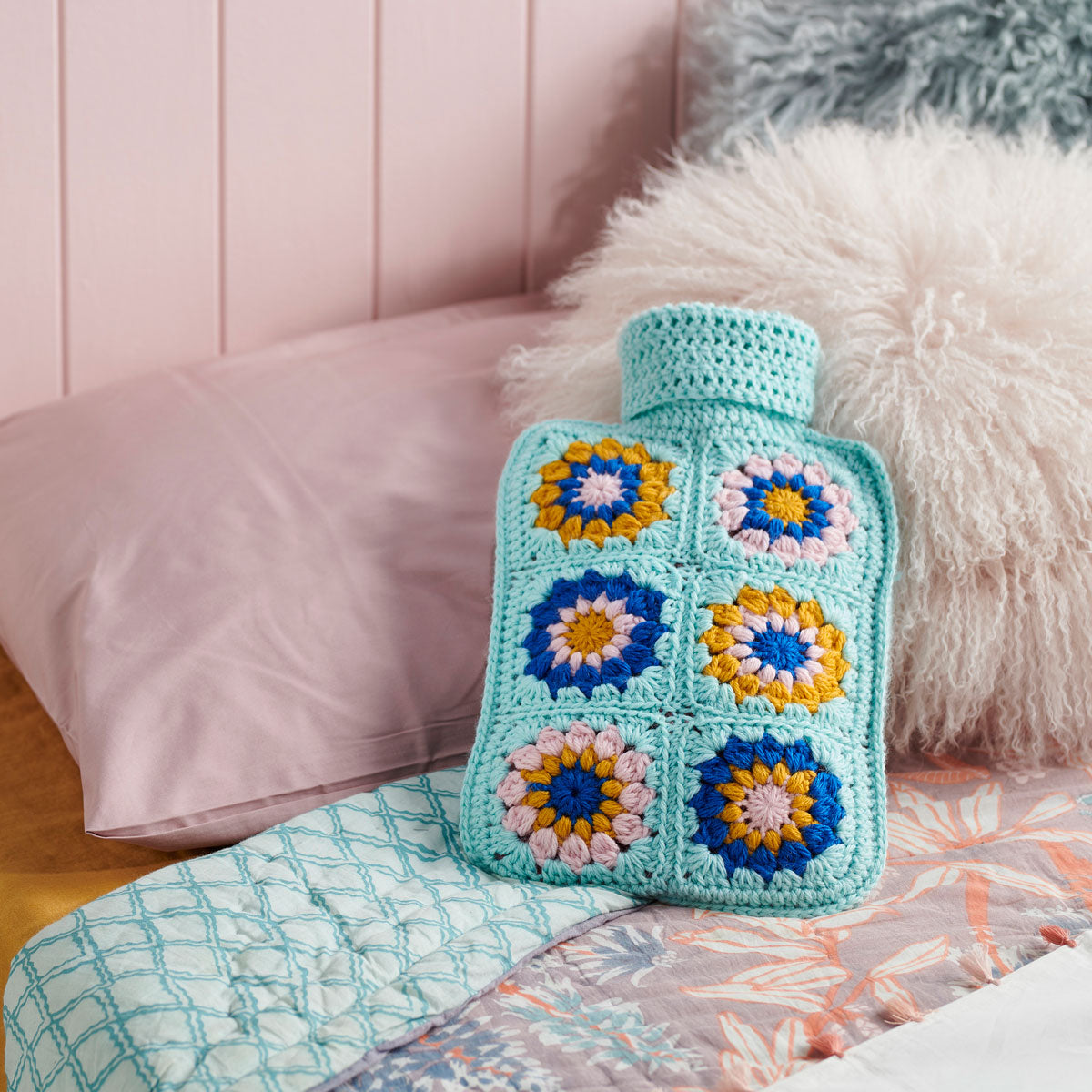 A crochet hot water bottle cover with a floral granny square pattern in blue, yellow, and pink colours lying on a bed. 