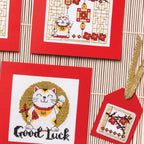 Cross-stitched designs featuring lucky cats on red fabric with a bamboo mat background.