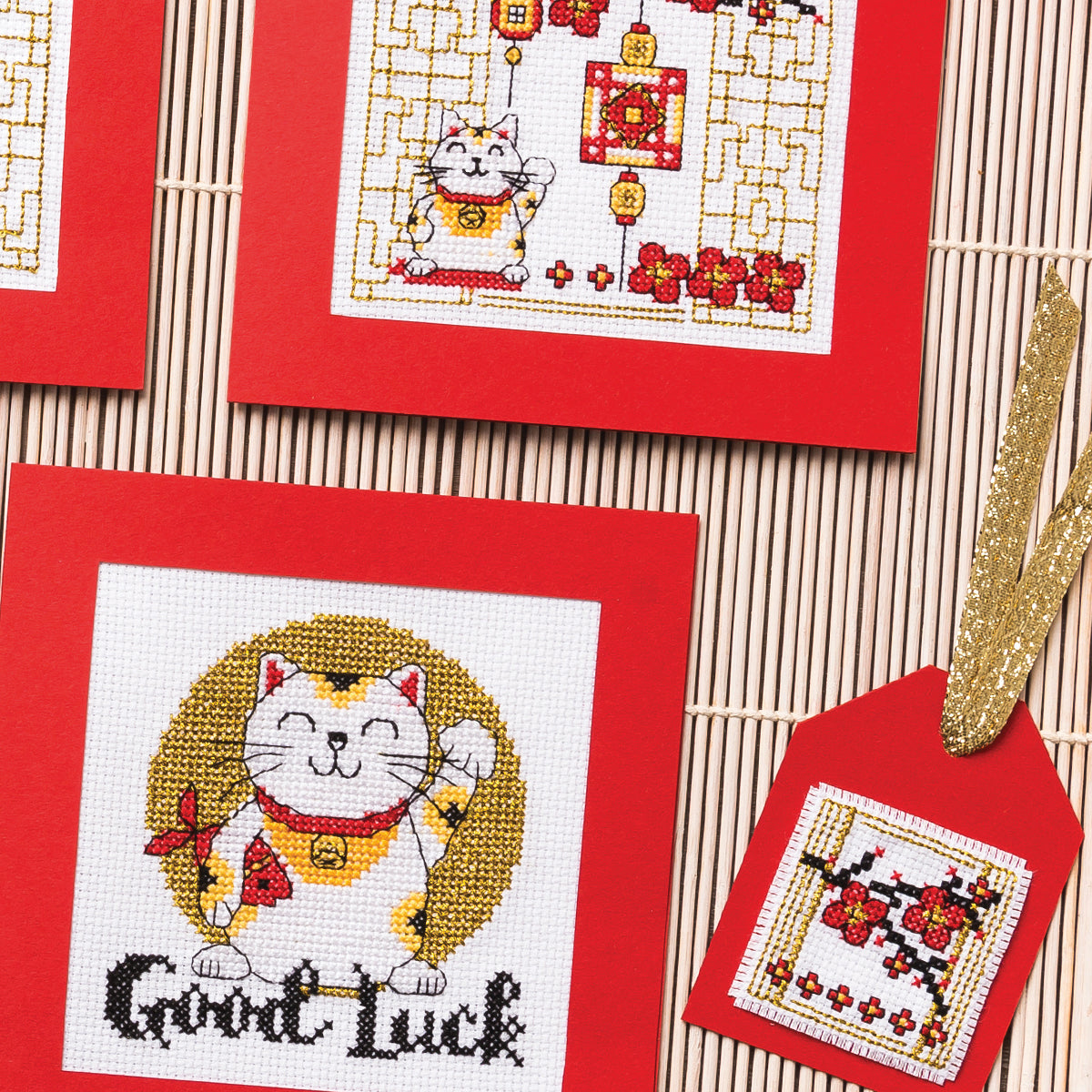 Cross-stitched designs featuring lucky cats on red fabric with a bamboo mat background.
