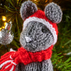 Knitted mouse toy wearing a red scarf and hat against a Christmas tree background