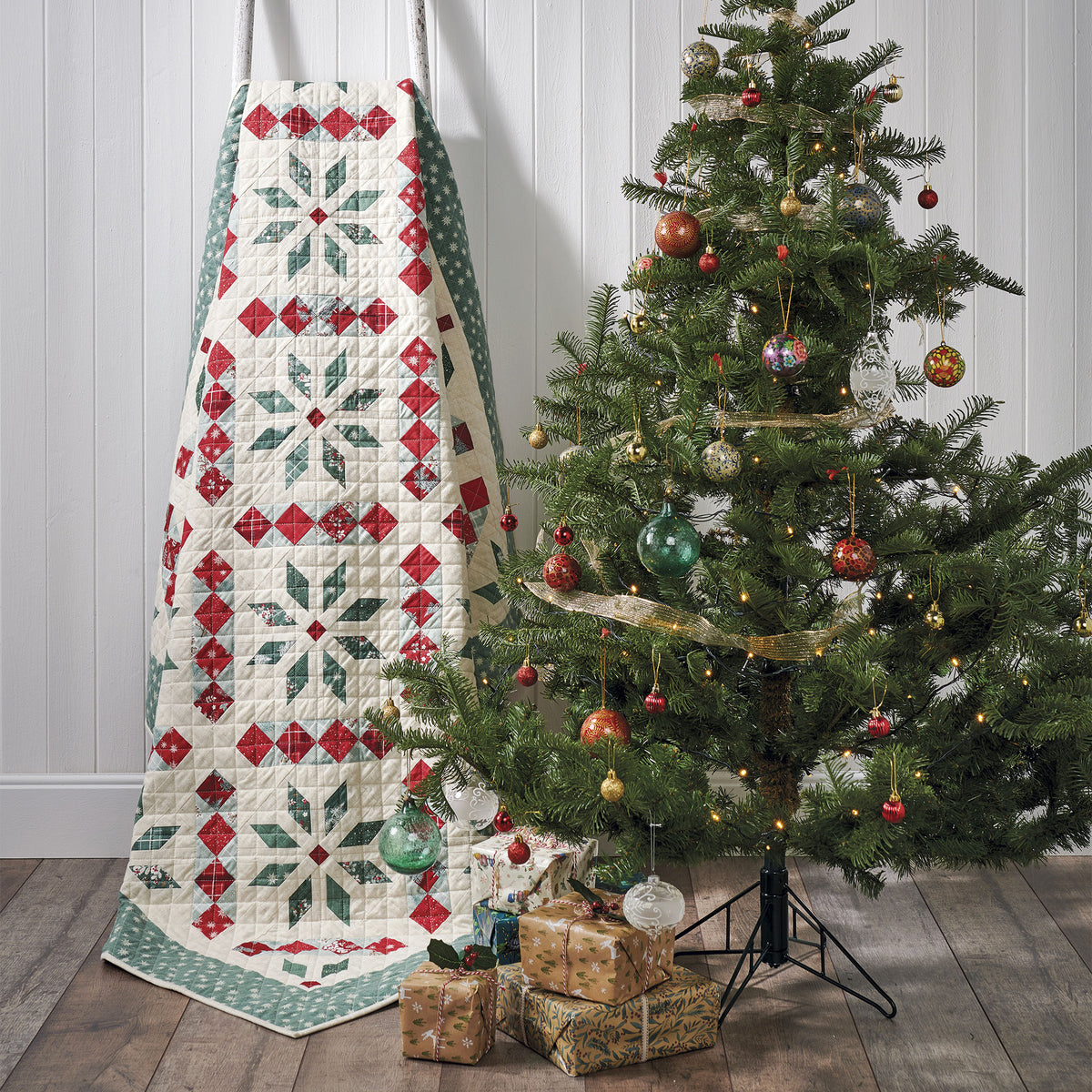 Decorative quilt with geometric pattern next to a Christmas tree with ornaments on a wooden floor.