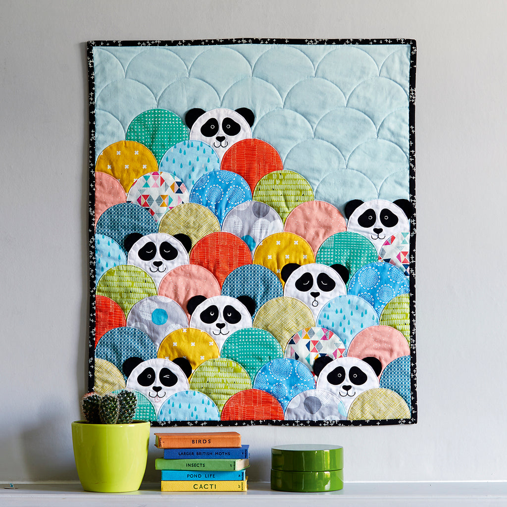 A colorful quilt featuring panda faces in a pattern with scallop edges, displayed on a wall with potted plants and books below.