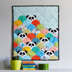A colorful quilt featuring panda faces in a pattern with scallop edges, displayed on a wall with potted plants and books below.